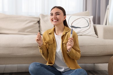 Happy woman holding pregnancy test on floor indoors