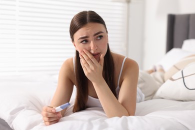 Sad woman holding pregnancy test on bed in room