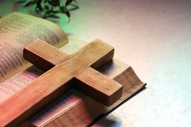 Photo of Wooden cross and Bible on textured table in color lights, closeup with space for text. Religion of Christianity