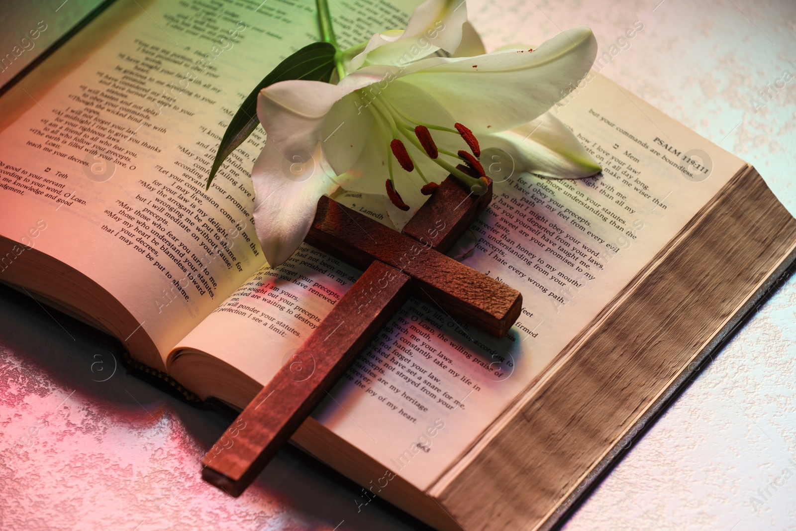 Photo of Wooden cross, Bible and lily flower on textured table in color lights, closeup. Religion of Christianity