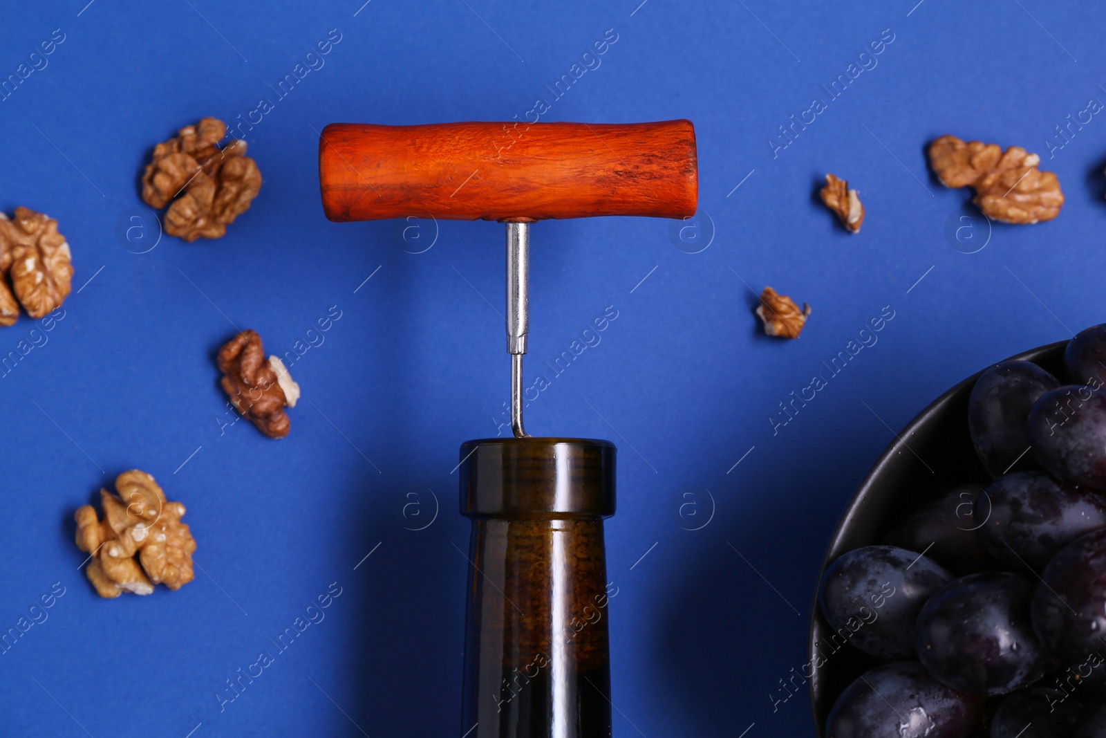 Photo of Bottle with corkscrew, grapes and walnuts on blue background, flat lay