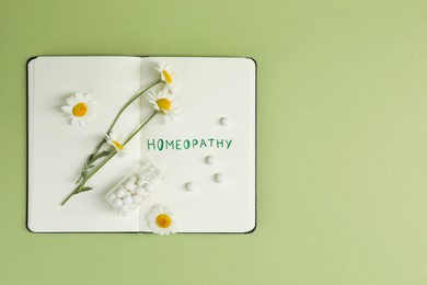 Photo of Notebook with word Homeopathy, pills and chamomile on green background, top view. Space for text