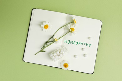 Photo of Notebook with word Homeopathy, pills and chamomile on green background, top view