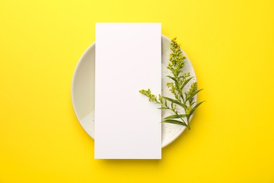 Empty menu, plate and floral decor on yellow background, top view. Mockup for design