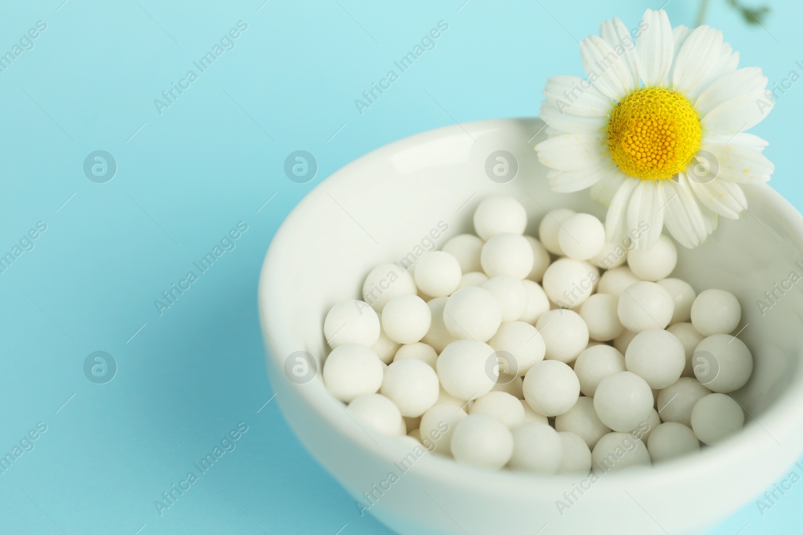 Photo of Homeopathy. Many pills and chamomile flower in bowl on light blue background, space for text