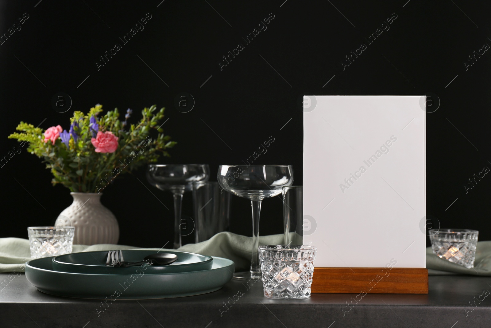 Photo of Menu holder, clean dishware and burning candles on grey table against black background