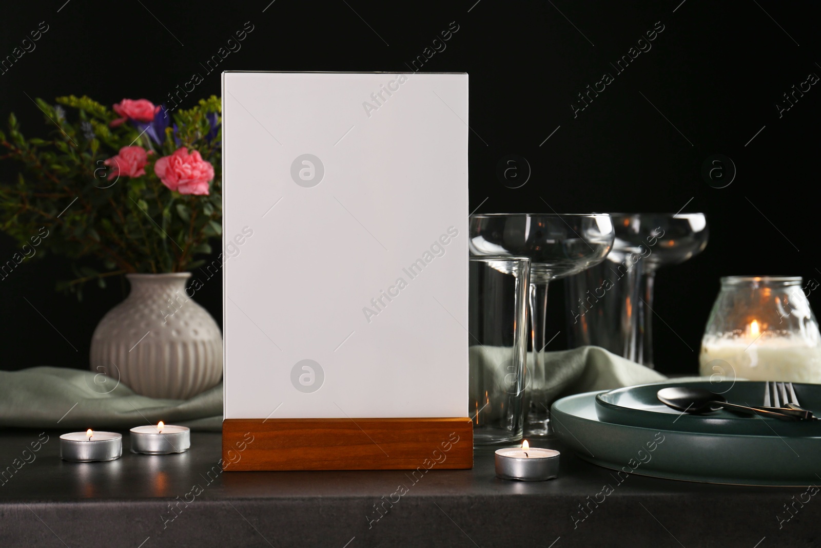 Photo of Menu holder, clean dishware and burning candles on grey table against black background