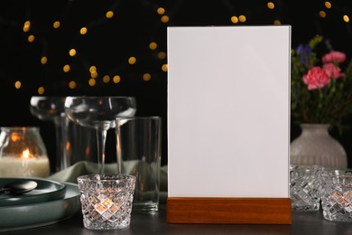 Photo of Menu holder, clean dishware and burning candles on grey table against blurred lights, closeup