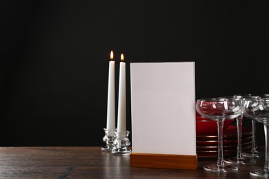 Photo of Menu holder, burning candles and clean dishware on wooden table against black background, space for text