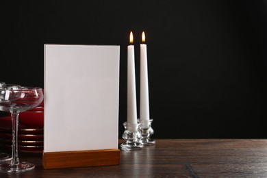 Photo of Menu holder, burning candles and clean dishware on wooden table against black background, space for text