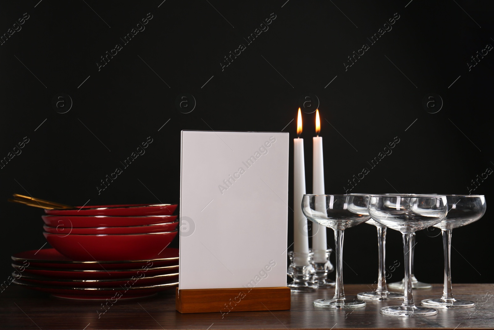 Photo of Menu holder, burning candles and clean dishware on wooden table against black background