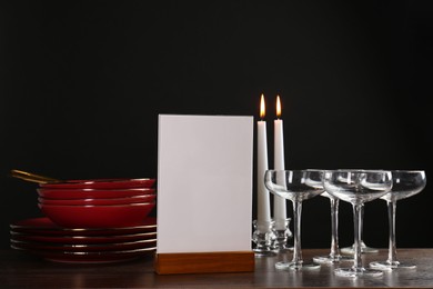 Photo of Menu holder, burning candles and clean dishware on wooden table against black background