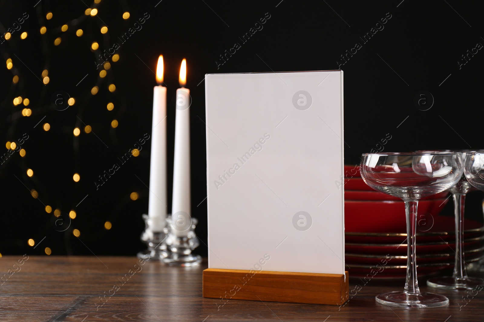 Photo of Menu holder, burning candles and clean dishware on wooden table against blurred lights