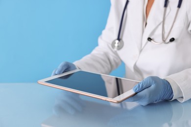 Doctor with tablet at table against light blue background, closeup view