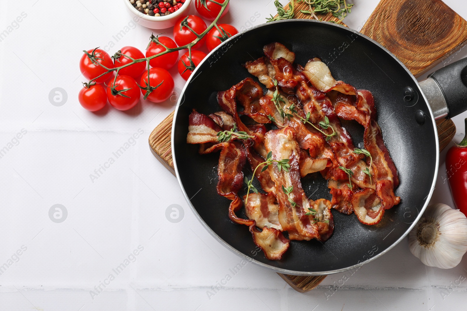 Photo of Delicious bacon slices in frying pan and products on white tiled table, flat lay. Space for text