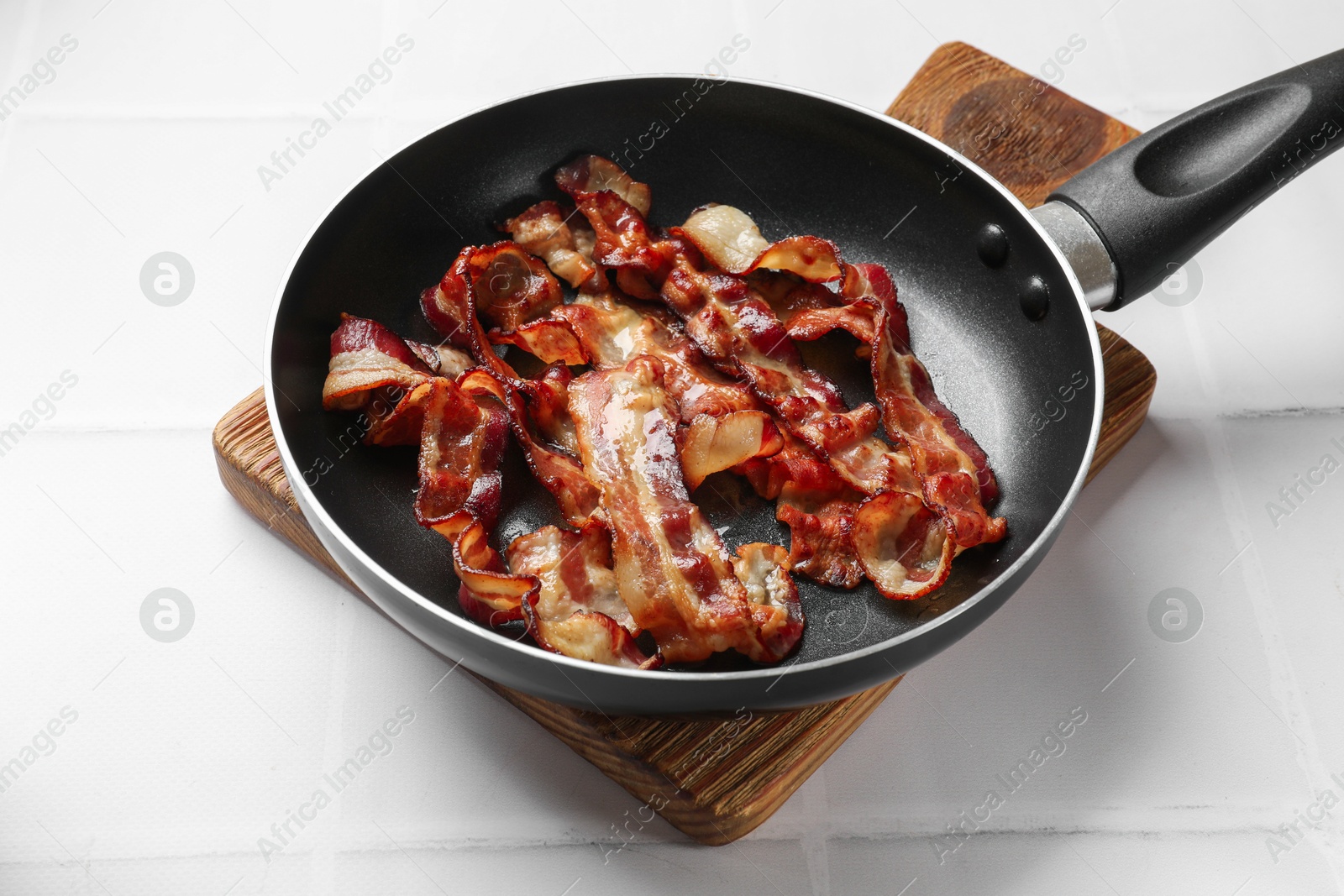 Photo of Delicious bacon slices in frying pan on white tiled table