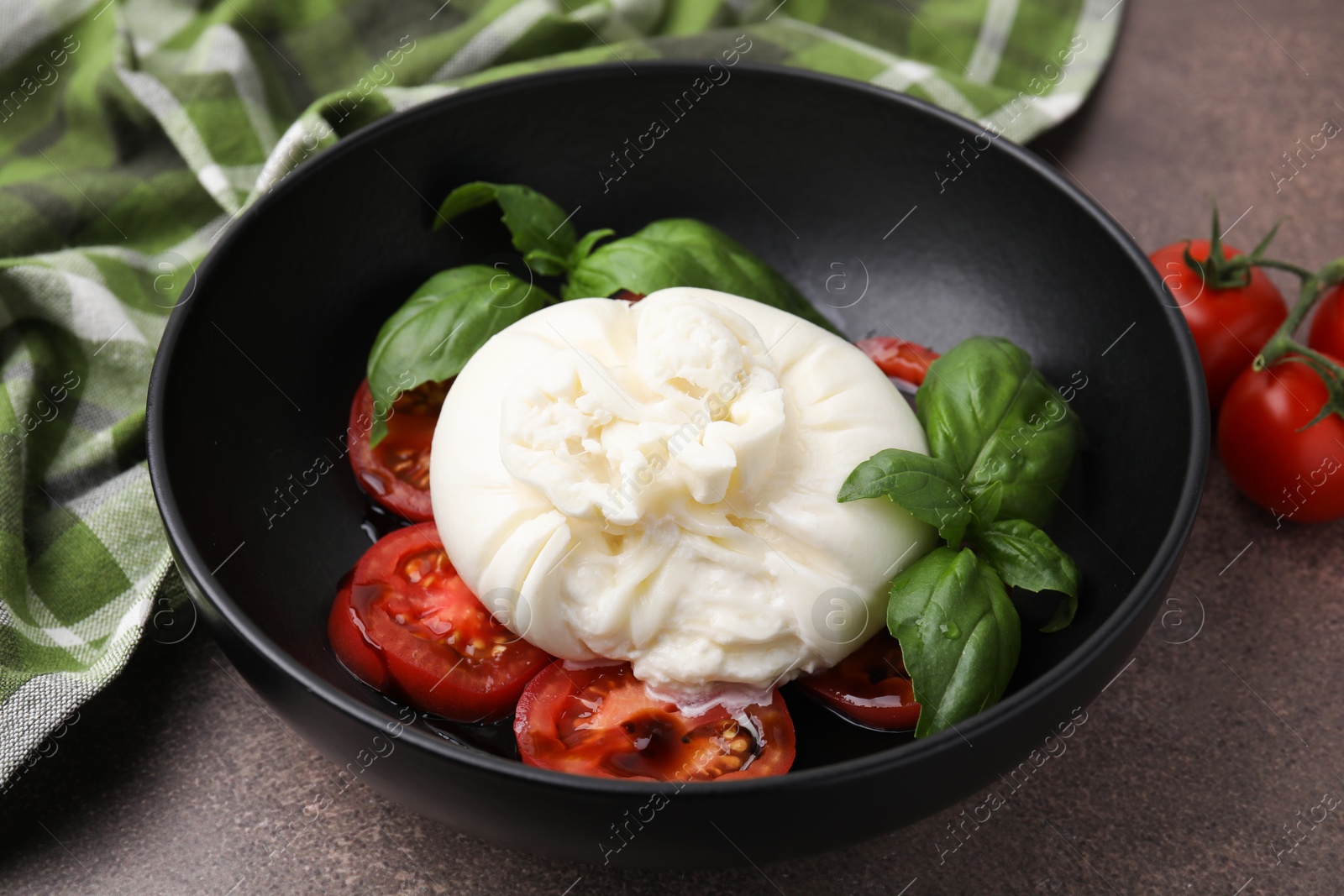 Photo of Delicious burrata cheese, tomatoes and basil in bowl on brown table, closeup