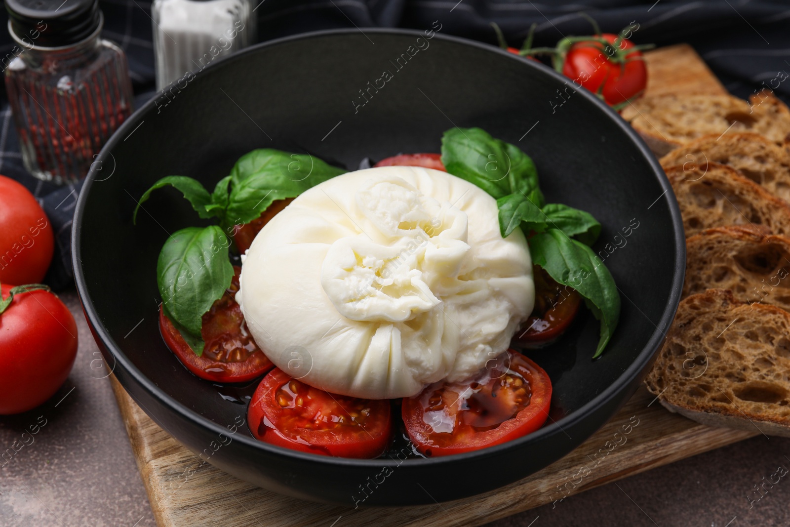 Photo of Delicious burrata cheese, tomatoes and basil in bowl on brown table, closeup