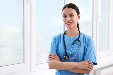 Photo of Smiling nurse with crossed arms near window in clinic. Space for text