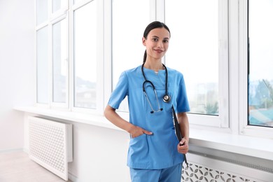 Beautiful nurse with clipboard near window in clinic. Space for text