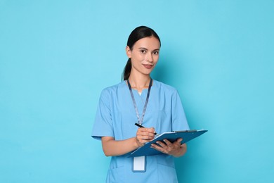 Photo of Beautiful nurse with clipboard writing notes on light blue background