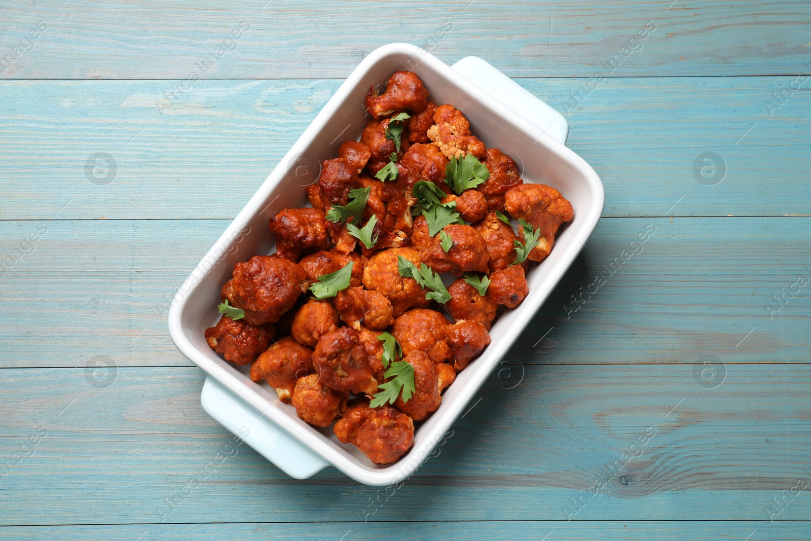 Photo of Baked cauliflower buffalo wings with parsley in baking dish on light blue wooden table, top view