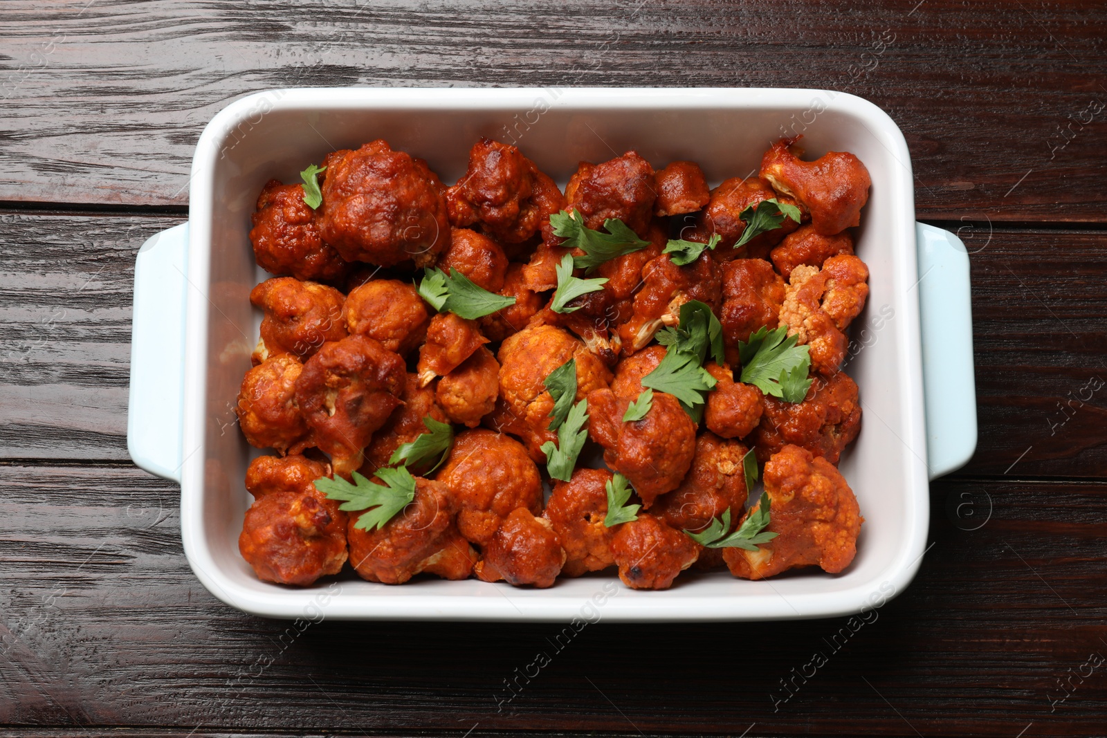 Photo of Baked cauliflower buffalo wings with parsley in baking dish on wooden table, top view