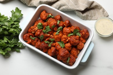 Photo of Baked cauliflower buffalo wings in baking dish, parsley and sauce on white marble table, top view