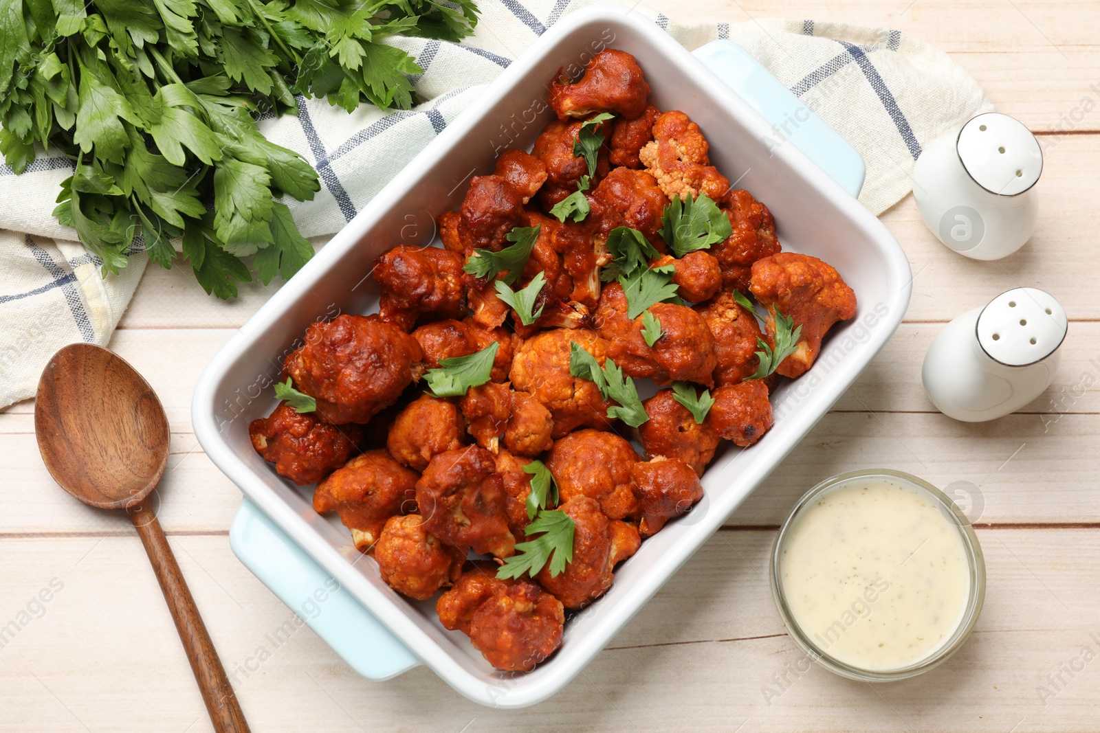 Photo of Baked cauliflower buffalo wings in baking dish, parsley, sauce and spoon on wooden table, top view