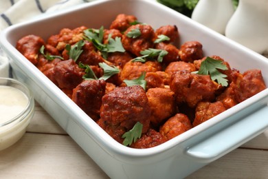 Photo of Baked cauliflower buffalo wings with parsley in baking dish and sauce on wooden table, closeup