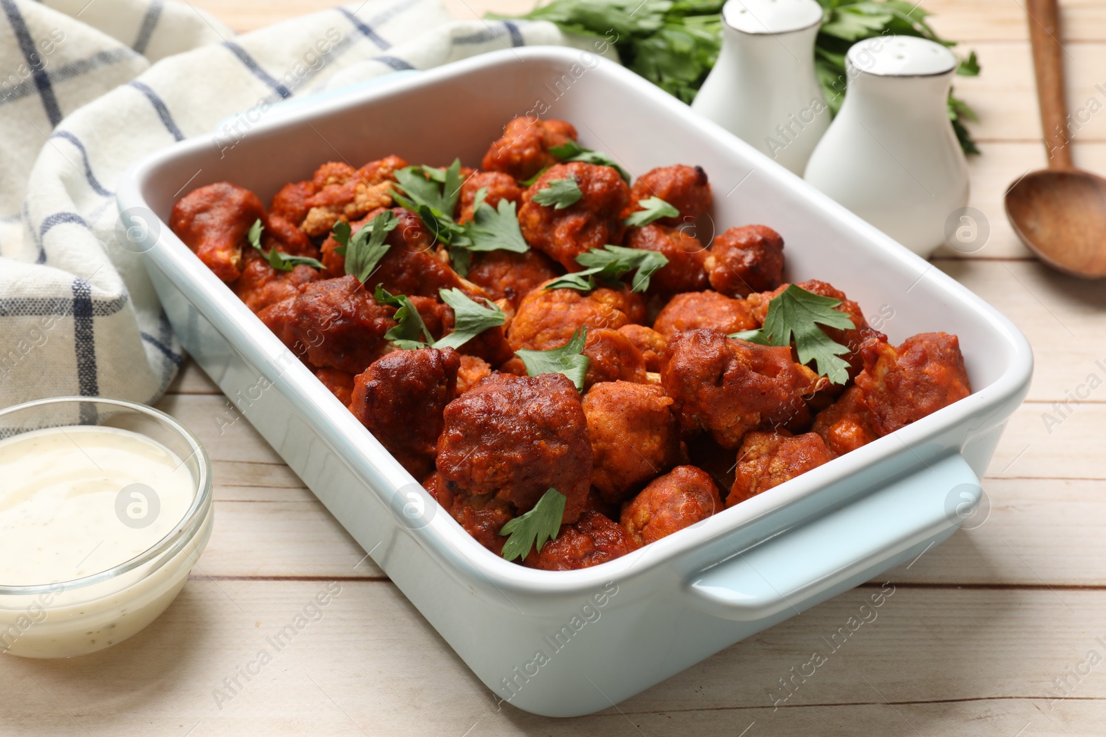 Photo of Baked cauliflower buffalo wings with parsley in baking dish and sauce on wooden table, closeup