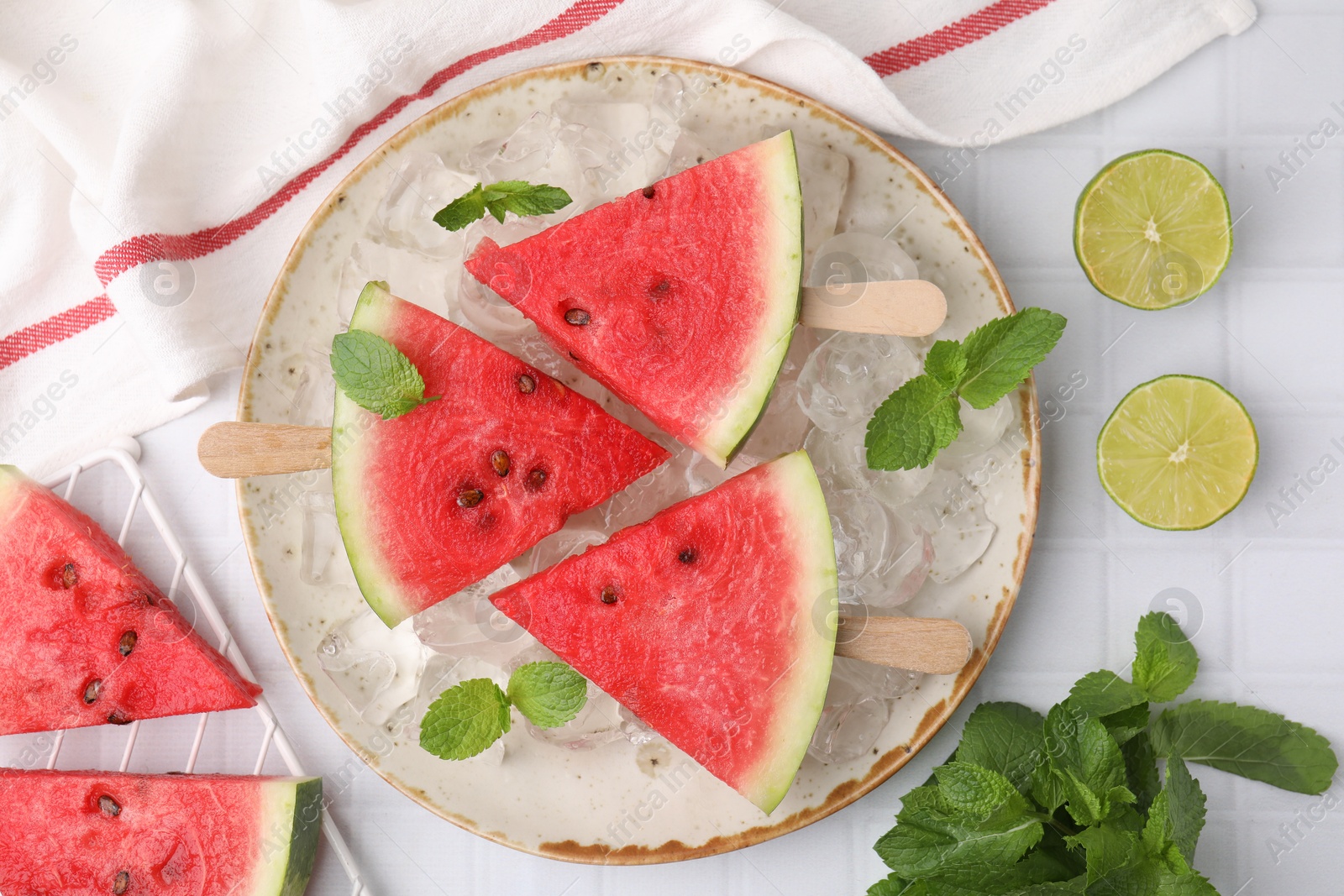 Photo of Pieces of tasty watermelon, ice cubes, lime and mint on white tiled table, flat lay