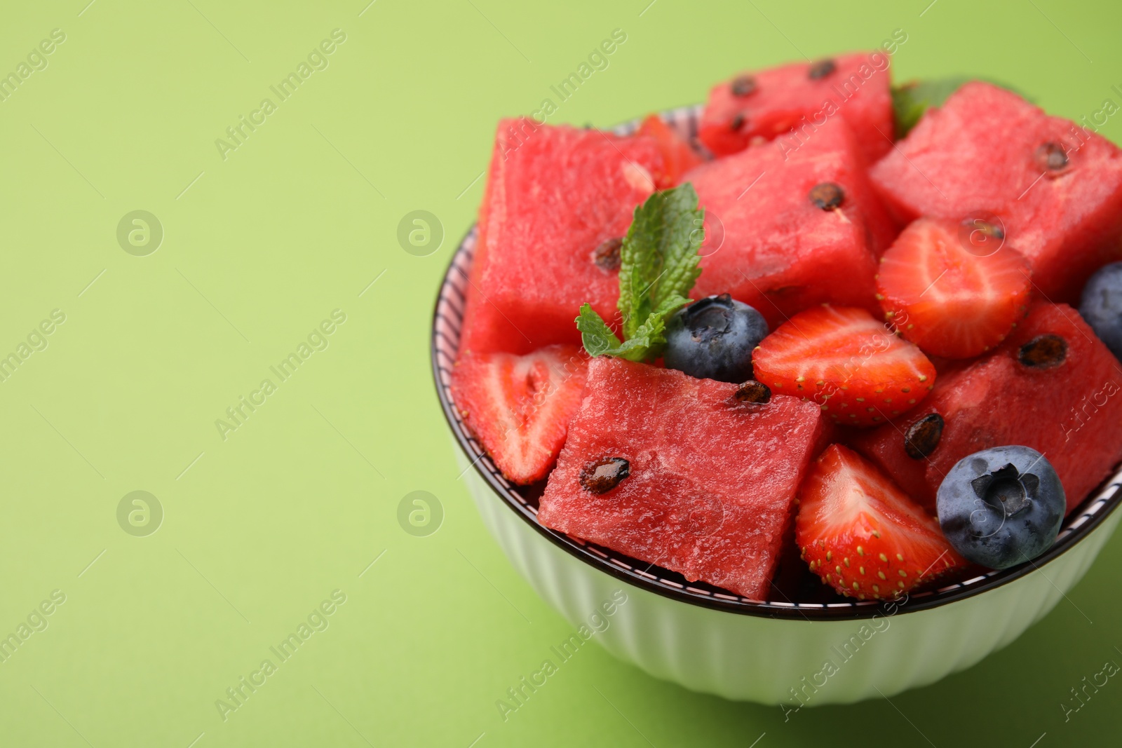 Photo of Pieces of tasty watermelon, strawberries, blueberries and mint in bowl on green table, closeup. Space for text