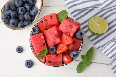 Photo of Pieces of tasty watermelon, strawberries, blueberries and mint in bowl on white wooden table, flat lay