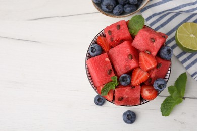 Photo of Pieces of tasty watermelon, strawberries, blueberries and mint in bowl on white wooden table, flat lay. Space for text