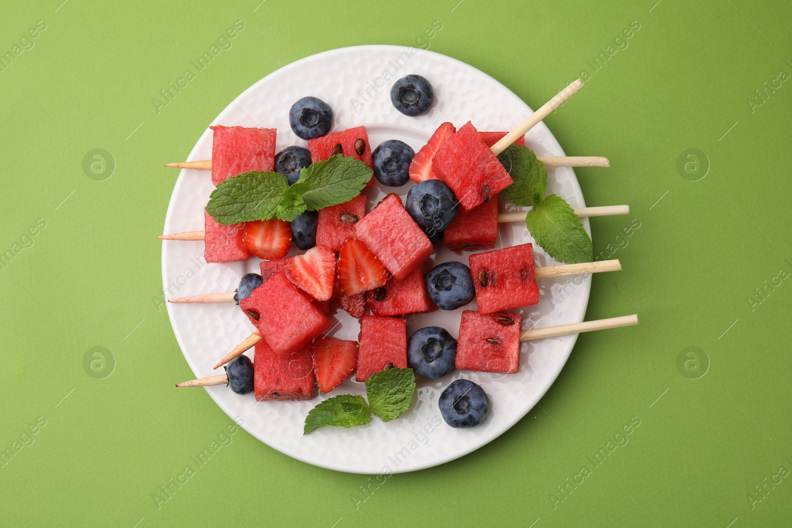 Photo of Skewers with tasty watermelon, strawberries, blueberries and mint on green table, top view