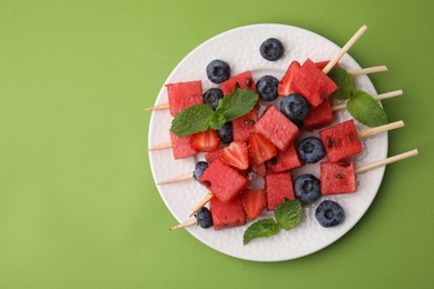 Photo of Skewers with tasty watermelon, strawberries, blueberries and mint on green table, top view. Space for text