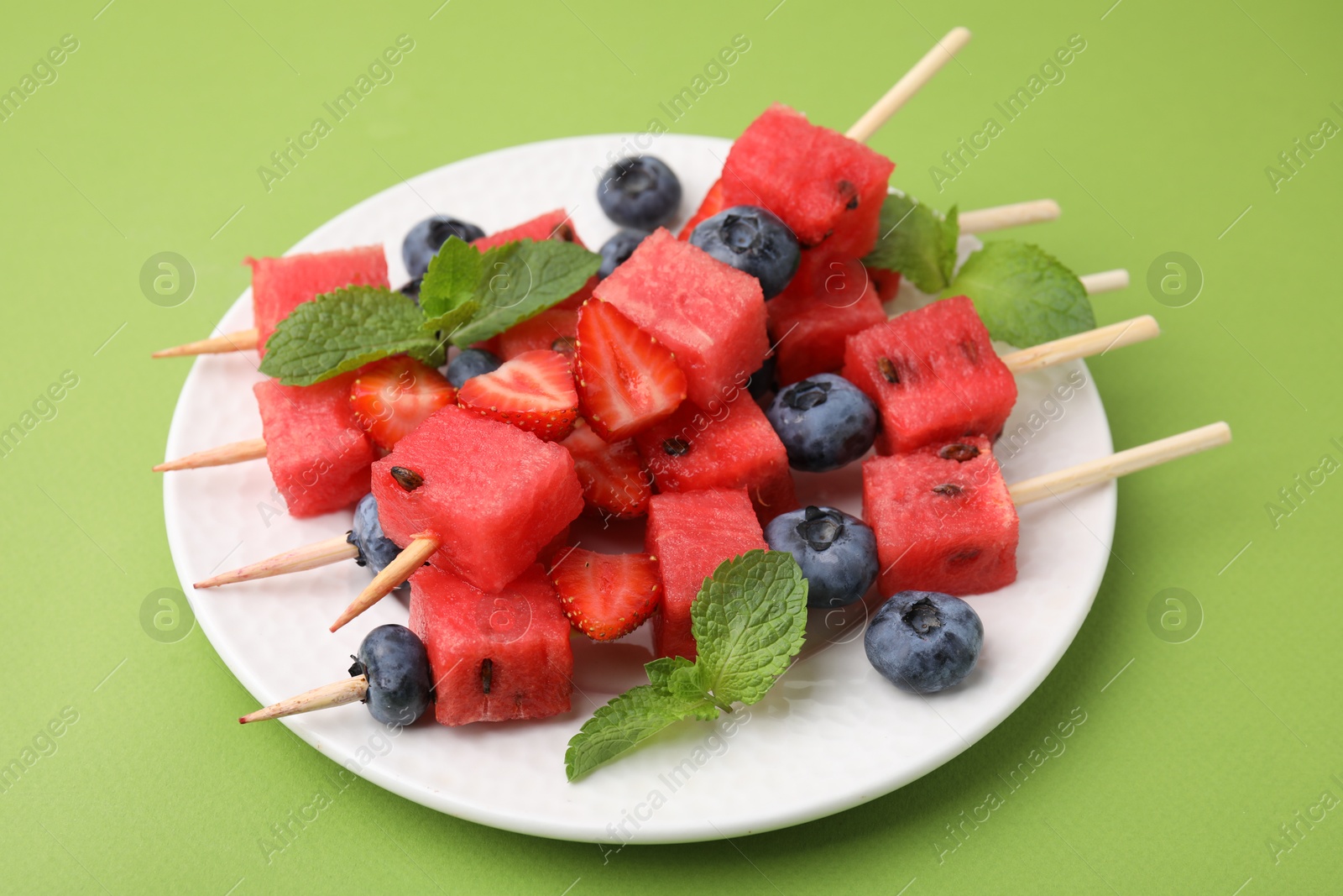 Photo of Skewers with tasty watermelon, strawberries, blueberries and mint on green table, closeup