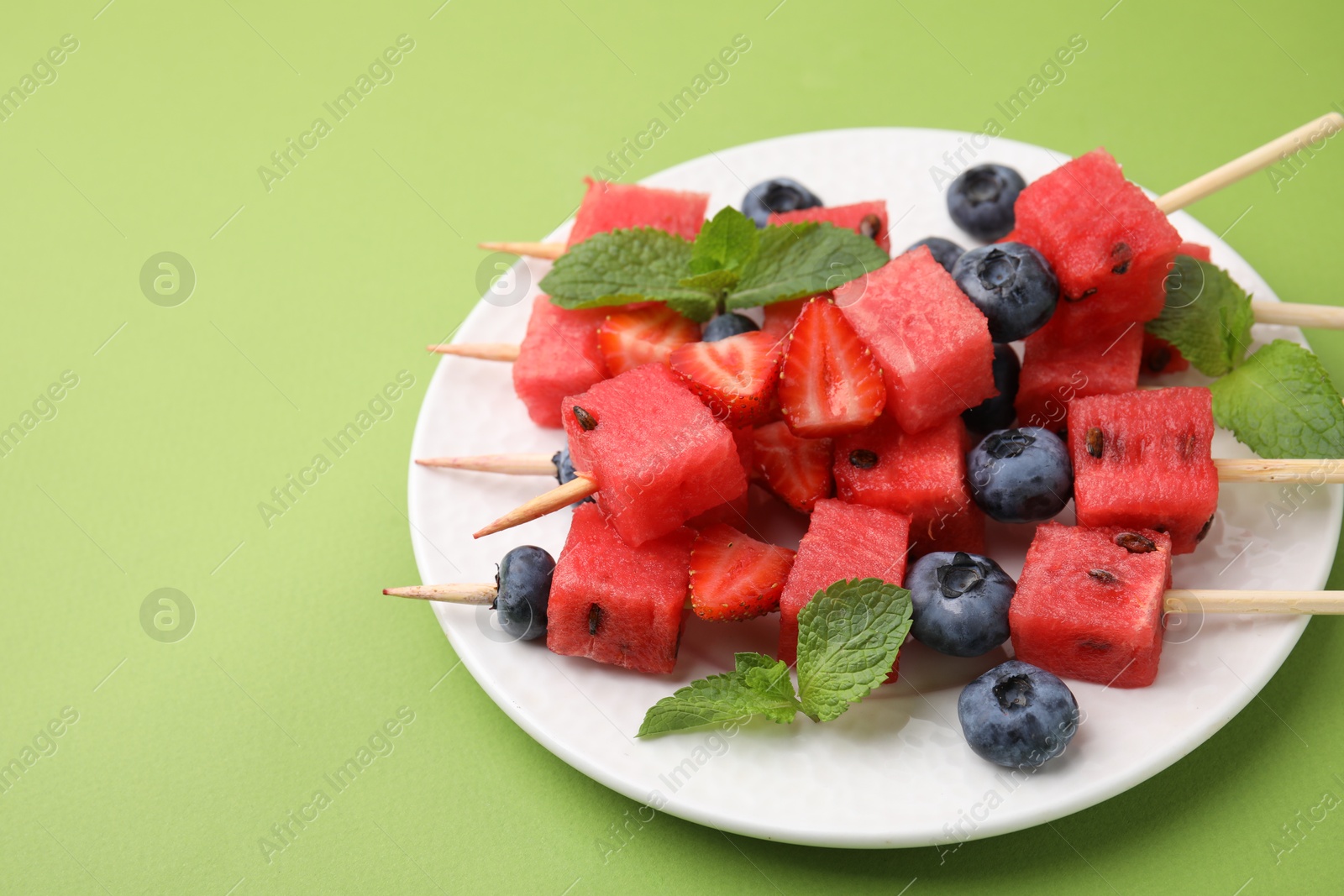 Photo of Skewers with tasty watermelon, strawberries, blueberries and mint on green table, closeup