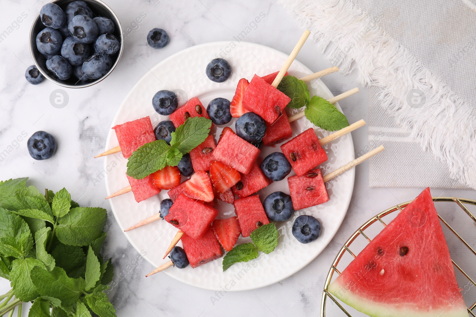 Photo of Skewers with tasty watermelon, strawberries, blueberries and mint on white marble table, flat lay