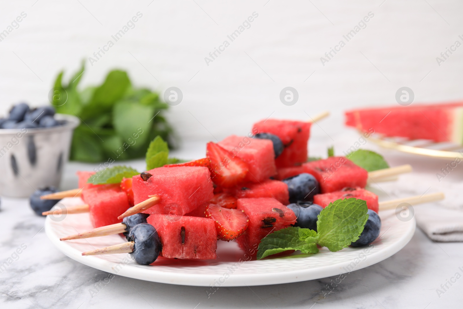 Photo of Skewers with tasty watermelon, strawberries, blueberries and mint on white marble table, closeup