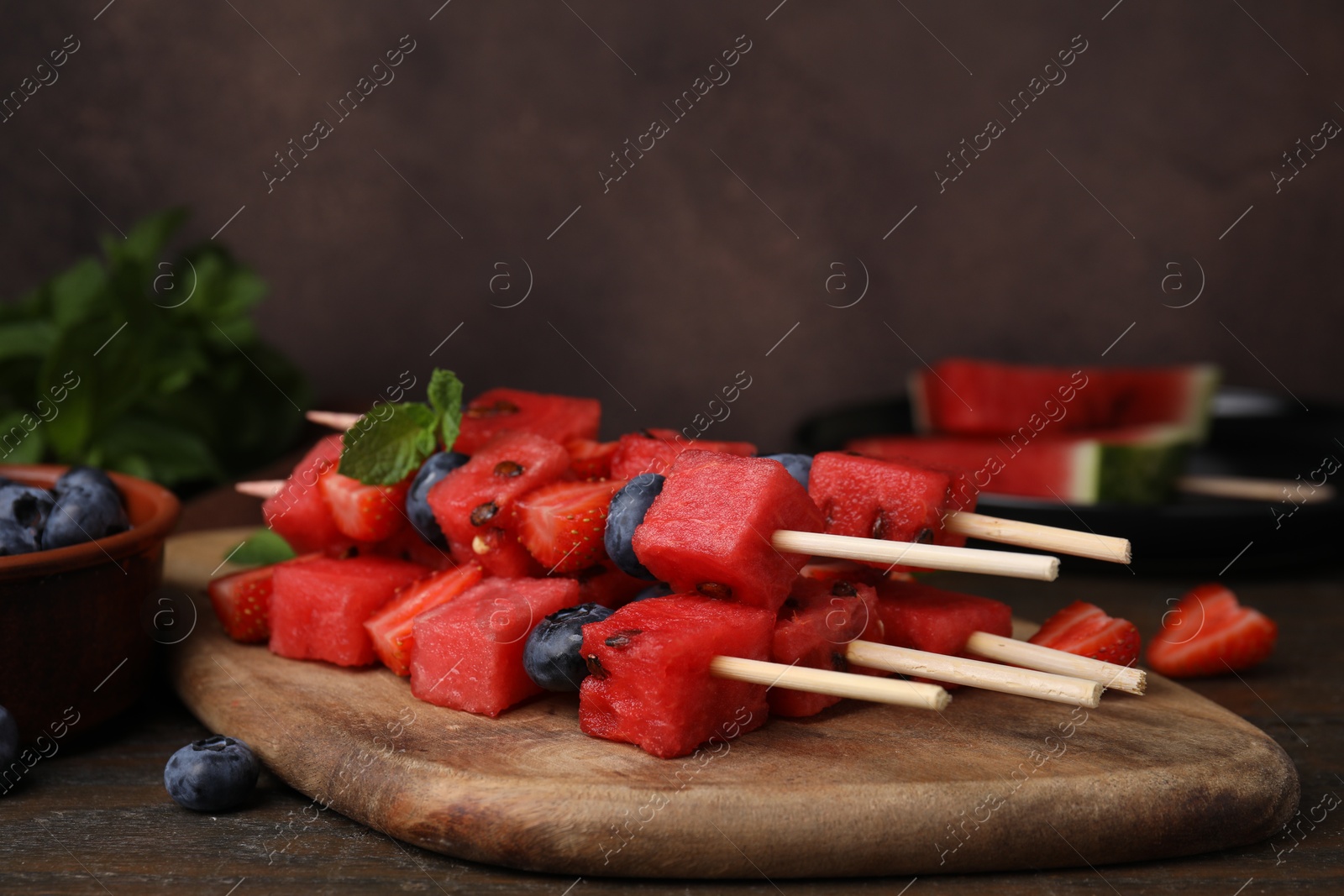 Photo of Skewers with watermelon, strawberries, blueberries and mint on wooden table against brown background, closeup