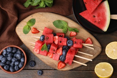 Photo of Skewers with tasty watermelon, strawberries, blueberries and mint on wooden table, flat lay