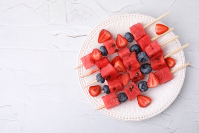 Photo of Skewers with tasty watermelon, strawberries and blueberries on white textured table, top view. Space for text