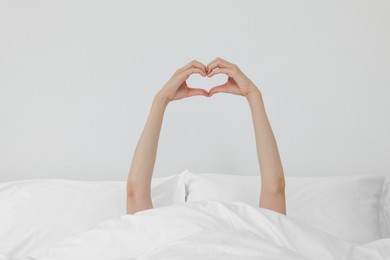 Bedtime. Woman showing heart gesture with hands in bed indoors, closeup