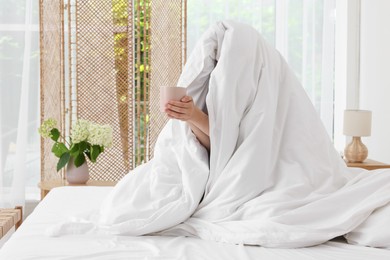 Photo of Bedtime. Woman wrapped in blanket holding cup of drink on bed indoors