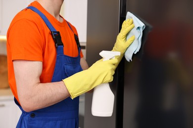 Photo of Professional janitor wearing uniform cleaning fridge in kitchen, closeup