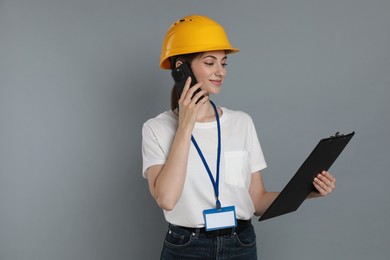 Engineer in hard hat with clipboard talking on smartphone against grey background, space for text