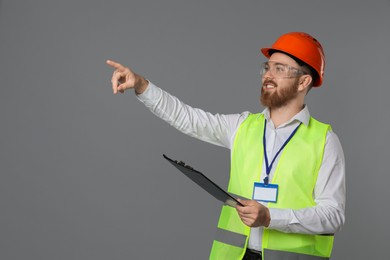 Engineer in hard hat and goggles holding clipboard on grey background, space for text