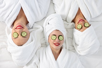 Photo of Happy friends in bathrobes with cucumber slices on bed, top view. Spa party